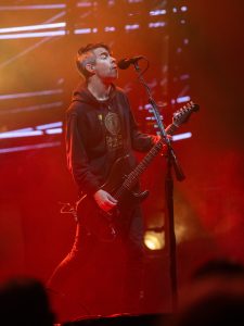 A man in a black hoodie playing a guitar in front of a red light.