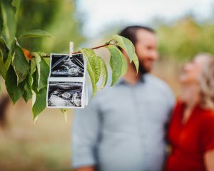 A pregnant couple is holding a photo of their baby on a tree.
