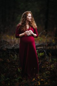A pregnant woman in a red dress standing in the woods.