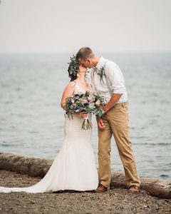 A bride and groom kiss on a log by the water.
