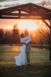 A bride and groom embrace in front of a gazebo at sunset.
